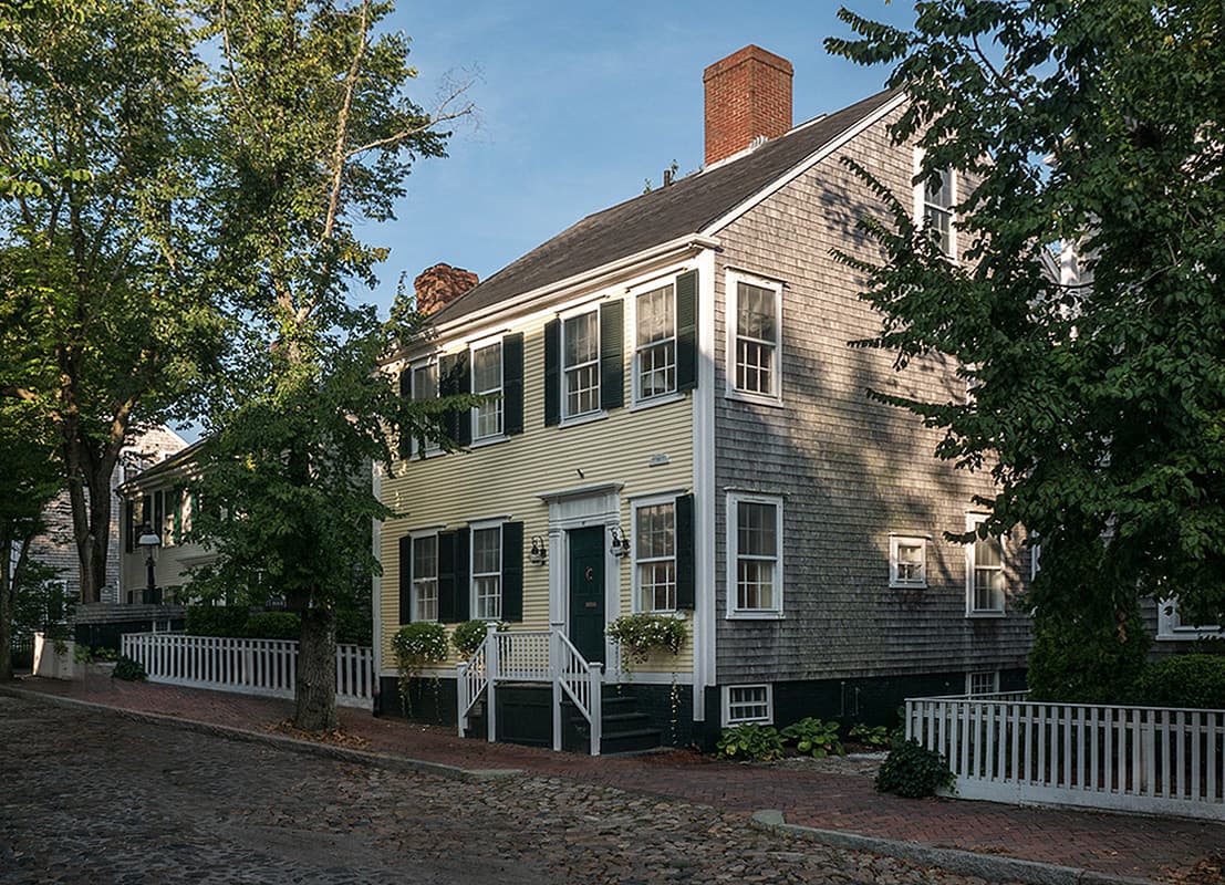 The house on its Main Street corner in daylight, yellow clapboard front and shingled flank above the cobblestones