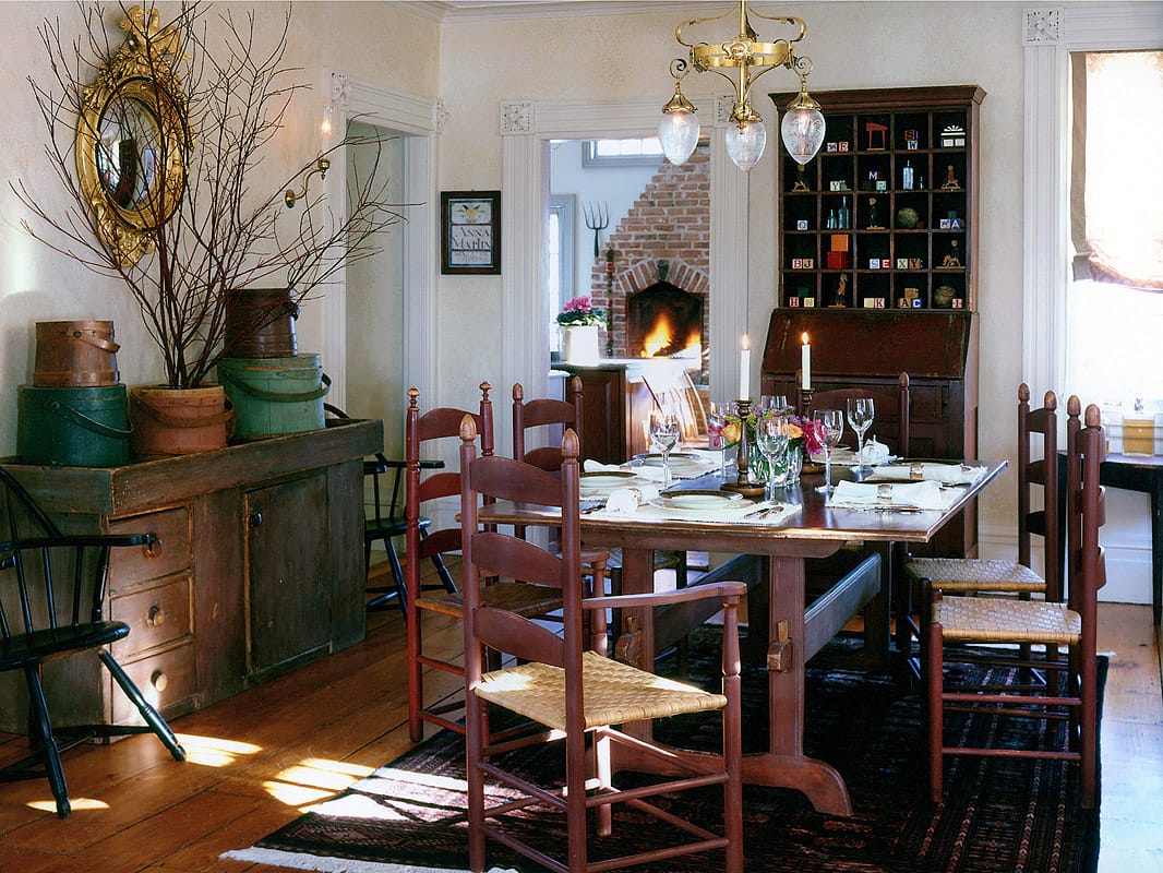 Dining room of the 1830 Federal house with a trestle table and red ladderback chairs, a bank of pigeonhole shelves and the kitchen fire beyond