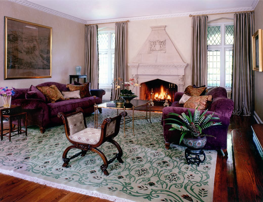 Living room of the 1925 Tudor with a carved limestone chimneypiece, plum velvet sofas either side of the lit fire, a curule stool on the scrolling green carpet and leaded casements on both walls