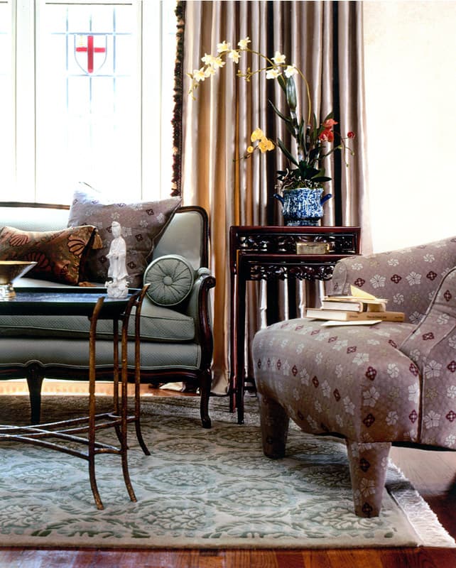 Bay window corner with a pale green settee under a stained glass crest, bamboo nesting tables, a carved Chinese stand holding an orchid and a patterned ottoman in the foreground