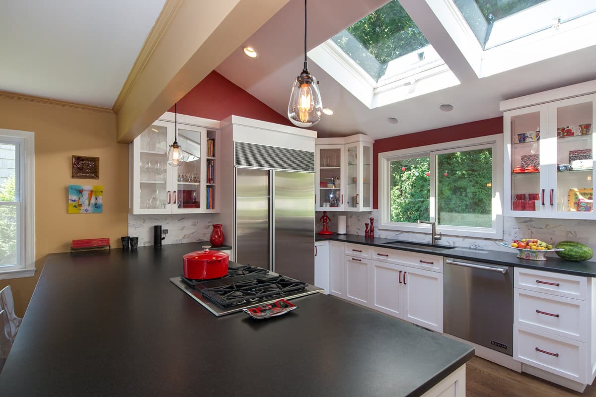 The kitchen from the island, two skylights in the vault above, a deep red gable wall, glass fronted upper cabinets and a stainless refrigerator set into white joinery