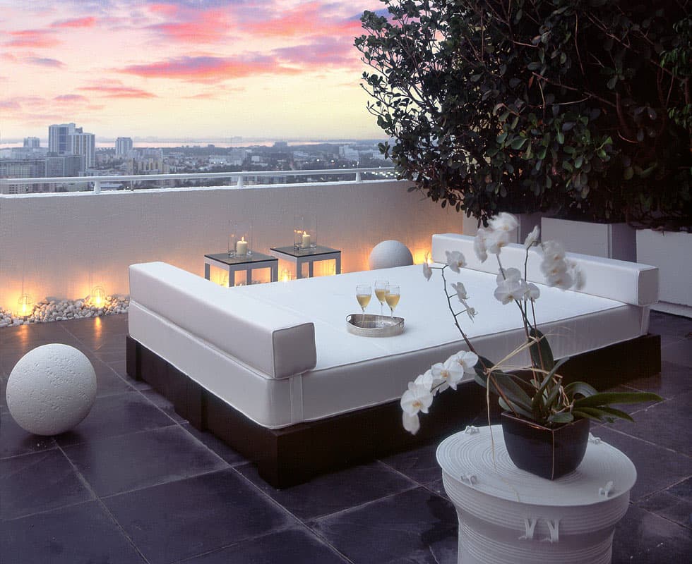 The Yacht Club Penthouse terrace at dusk, a white daybed and lanterns on dark stone above the Miami Beach skyline