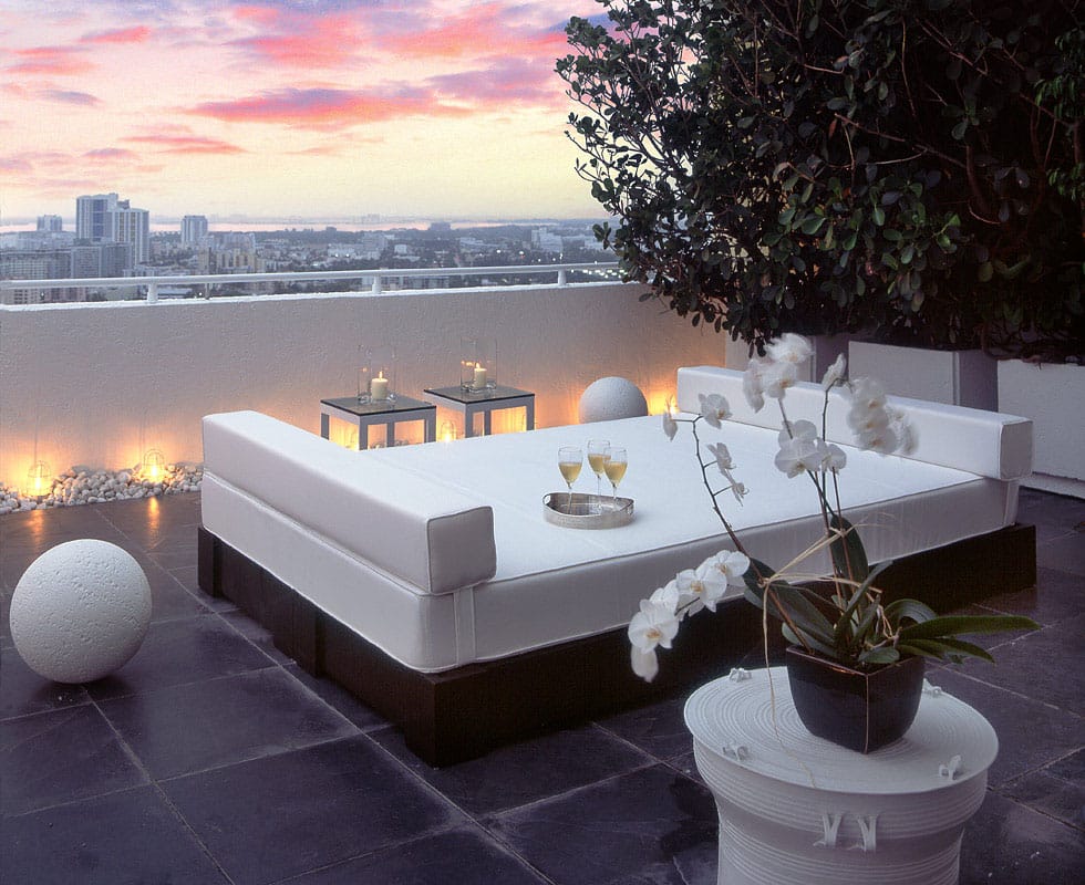 The penthouse terrace at dusk, a wide white daybed and lanterns on dark stone paving above the Miami Beach skyline