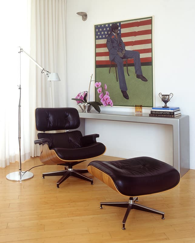 A black leather and plywood lounge chair with its ottoman at a grey console, a framed painting of a seated figure against a flag above, orchids and a stack of books alongside