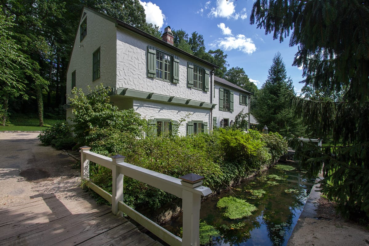 The white stucco Cotswold cottage with green shutters seen across a timber footbridge over the mill stream, spruce and shrubbery crowding the water