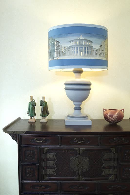 A pale urn lamp with a shade painted with a domed temple and city street, standing on a dark Korean chest with iron hardware between two green glazed figures and a pink pottery bowl