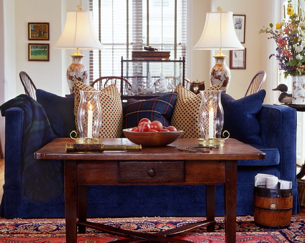 Blue slipcovered sofa with a tartan throw, lattice cushions and a bowl of cricket balls on a scrubbed table in front, hurricane candles lit, a pair of Chinese vase lamps and Windsor chairs beyond