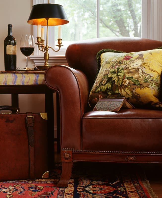 Brown leather club chair with a yellow needlework pheasant cushion, a brass bouillotte lamp with a black shade, a glass of red wine and a strapped leather case at the chair side