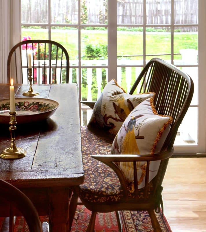 Scrubbed farm table with brass candlesticks and a painted bowl, Windsor chairs and a window bench cushioned in a small blue print, French doors to the garden behind