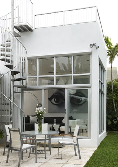 The white stucco house from the garden, a steel spiral stair to the roof deck, a glazed corner showing the eye paintings inside and a terrace table on pale paving
