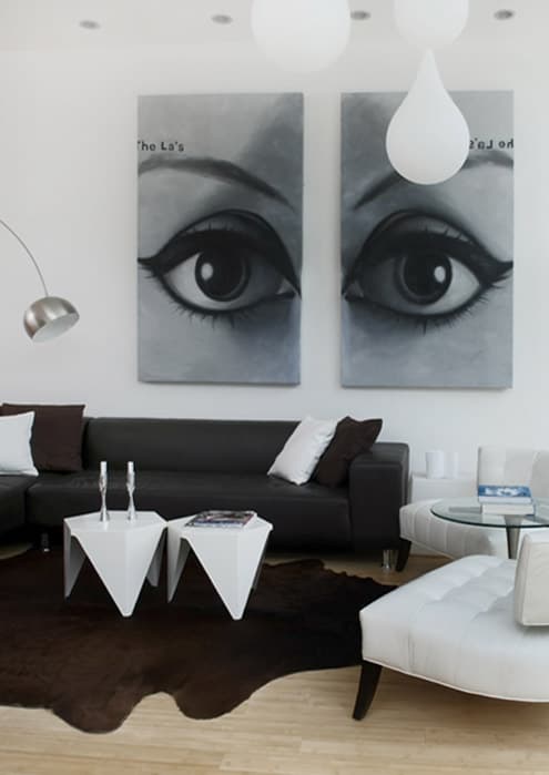 Living room with a grey diptych of a woman's eyes above a dark leather sectional, two faceted white side tables, a white tufted chaise and a cowhide on the bamboo floor