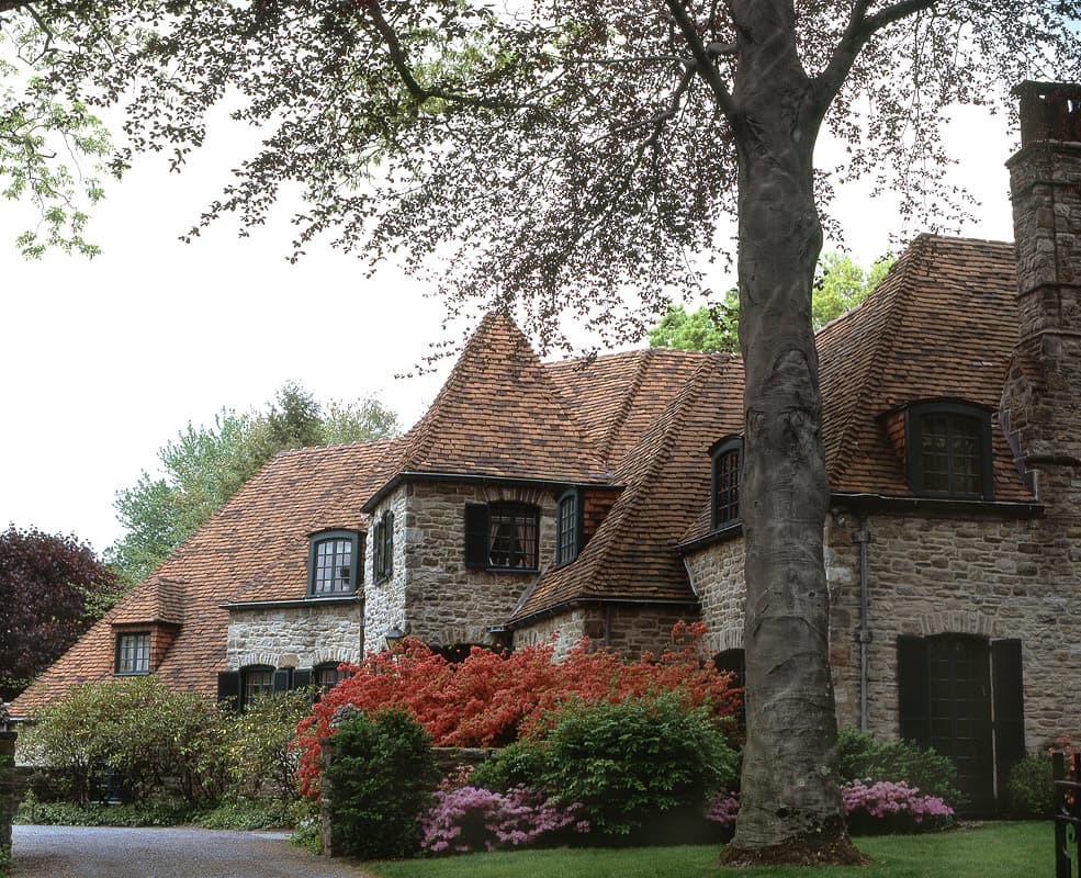 The French Normandie Tudor in New Rochelle, stone under a steep clay tile roof with a copper beech and banks of azalea