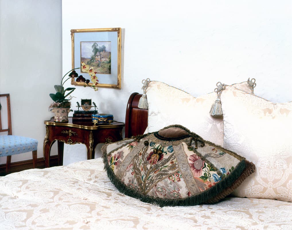 An embroidered silk fan shaped cushion with green fringe lying on a cream damask bed, tasselled shams behind and a marquetry bedside table with an orchid and a framed landscape above