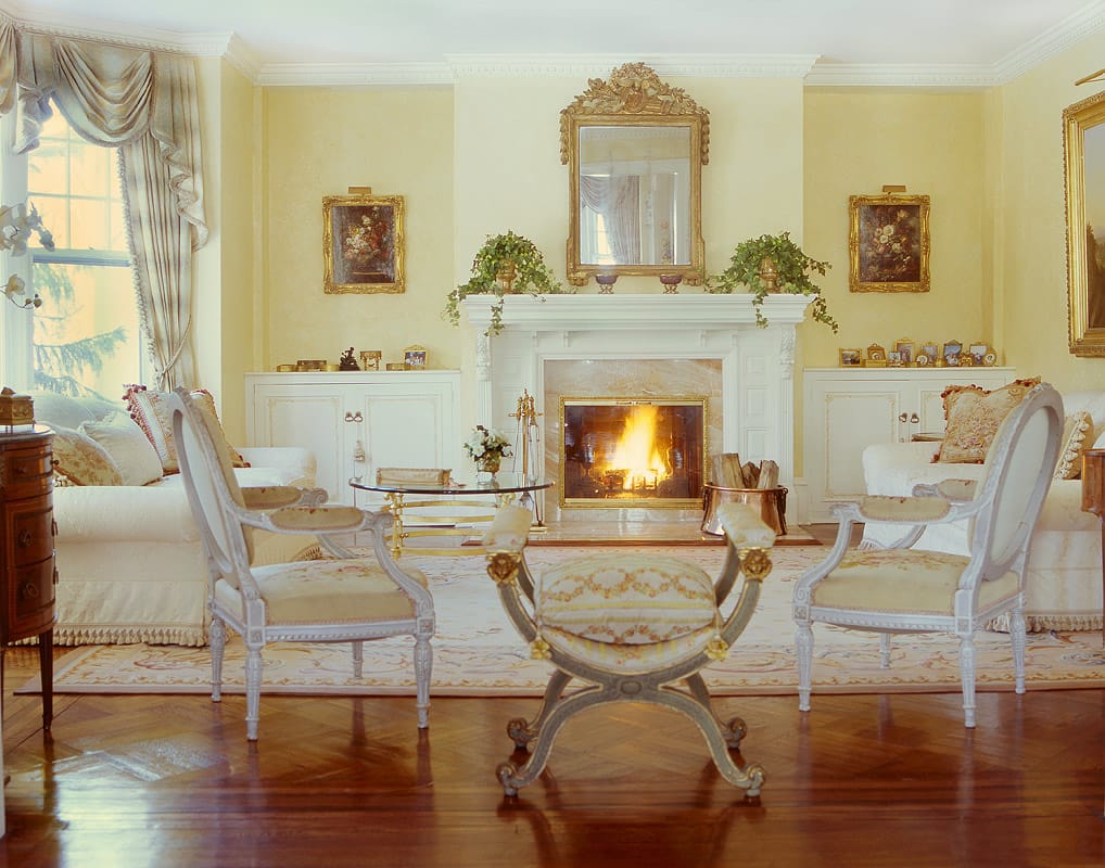 Living room with a white marble chimneypiece alight, a carved gilt mirror above, cream slipcovered seating, painted Louis XVI armchairs and a curule stool on an Aubusson carpet