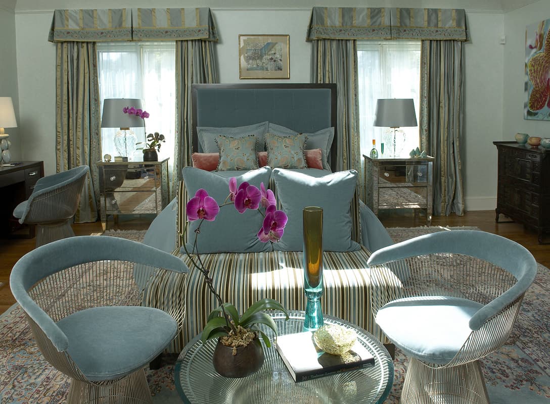 Master bedroom seen between two pale blue velvet lounge chairs, a tufted velvet bed under shaped silk valances with mirrored chests either side and orchids on the glass table