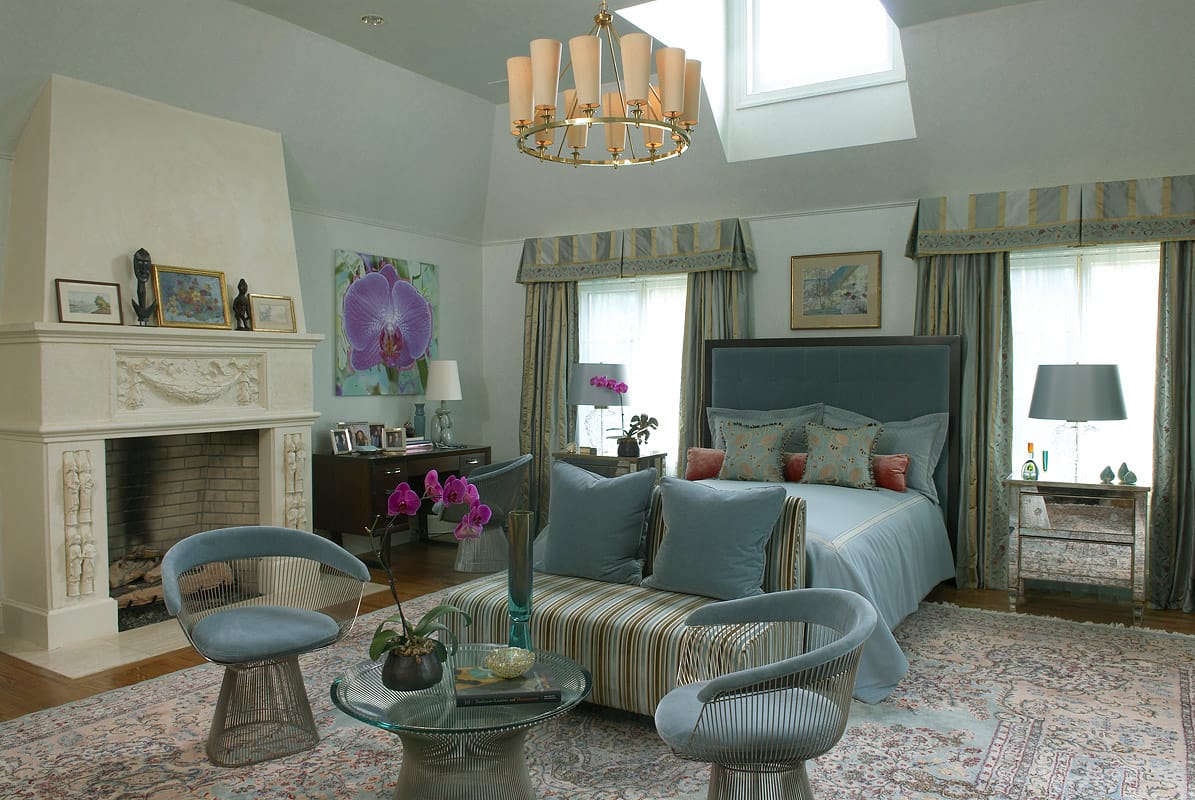 Master bedroom under a vaulted ceiling and skylight, a brass chandelier over a striped ottoman, a limestone chimneypiece to the left and the blue velvet bed between valanced windows