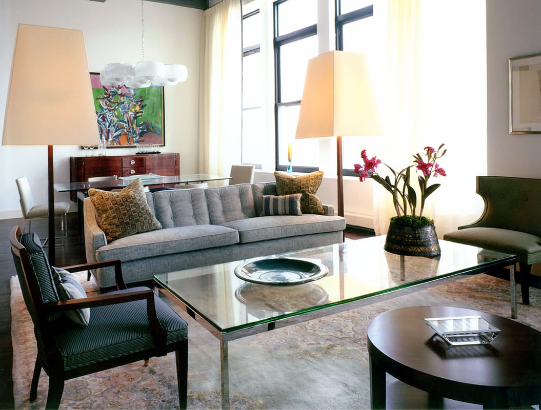 Loft living room with a grey flannel sofa between two tall shaded floor lamps, a chrome and glass table on a faded carpet and factory windows behind sheer curtains