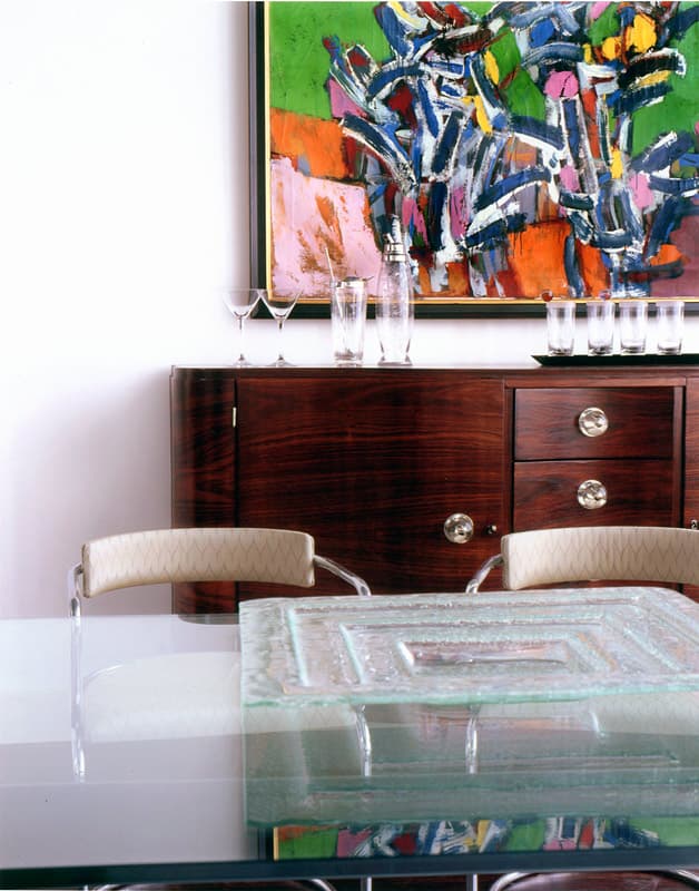Art Deco rosewood sideboard set with bar glass under a large green and blue abstract painting, lucite framed chairs and a thick cast glass box on the glass dining table in front