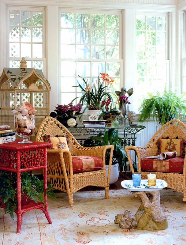 Sun porch with two natural wicker armchairs in red check cushions, a red painted wicker side table, a domed birdcage, a glass jar of shells, ferns and orchids at three tall windows