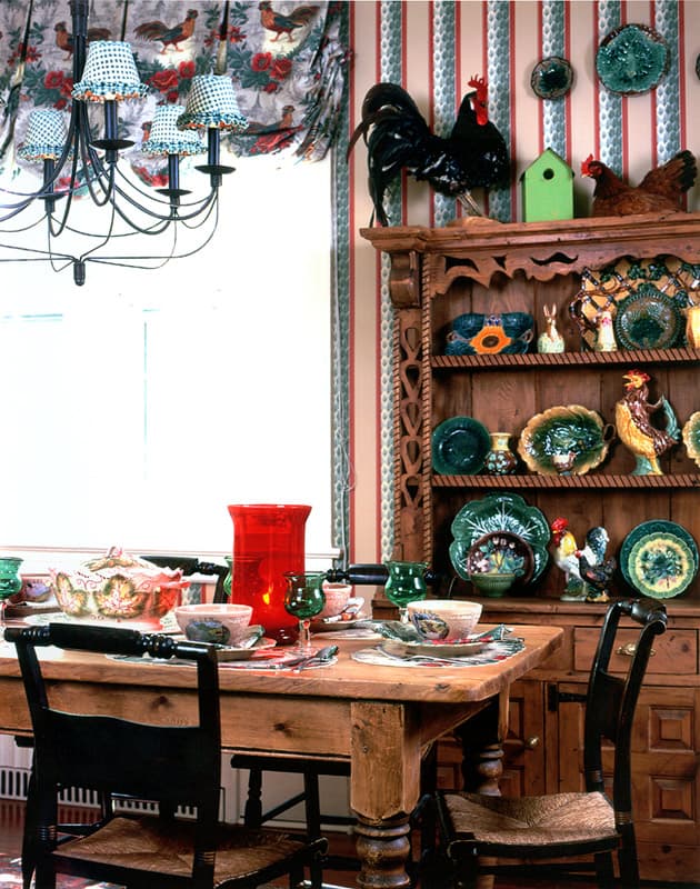 Kitchen dresser in pine stacked with green majolica, pottery roosters above, a scrubbed table laid with a red hurricane candle and painted china, black chairs and a rooster print valance at the window