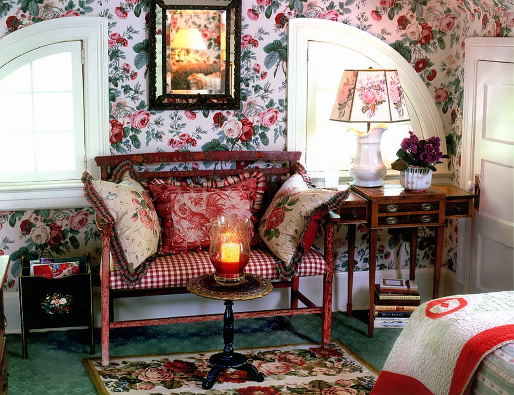 Bedroom corner papered in full scale roses, a red painted bench piled with toile and gingham cushions between two arched dormer windows, a jug and violets on an inlaid side table and a red and white quilt on the bed