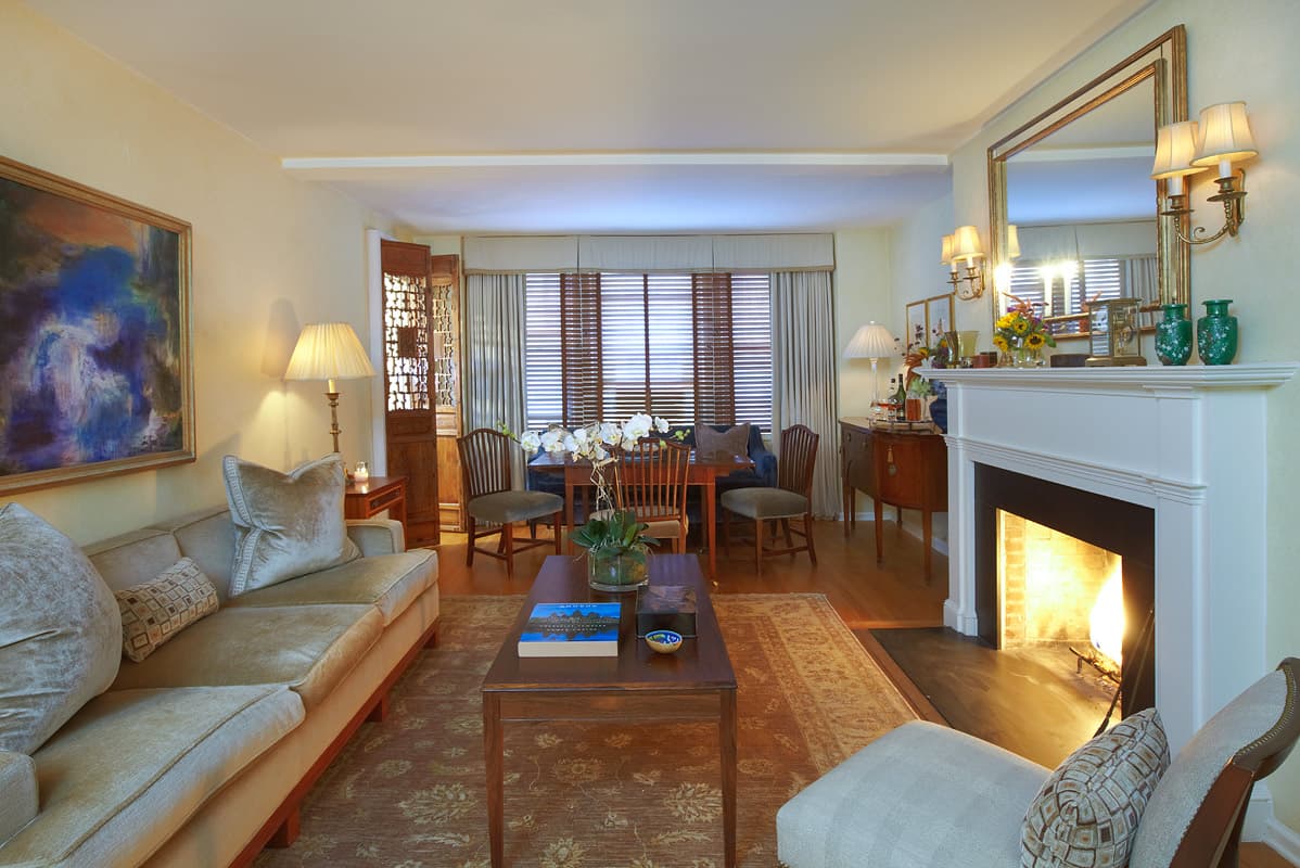 West Village living room with a lit fire under a white mantel, a gilt overmantel mirror and brass sconces and the dining bay beyond