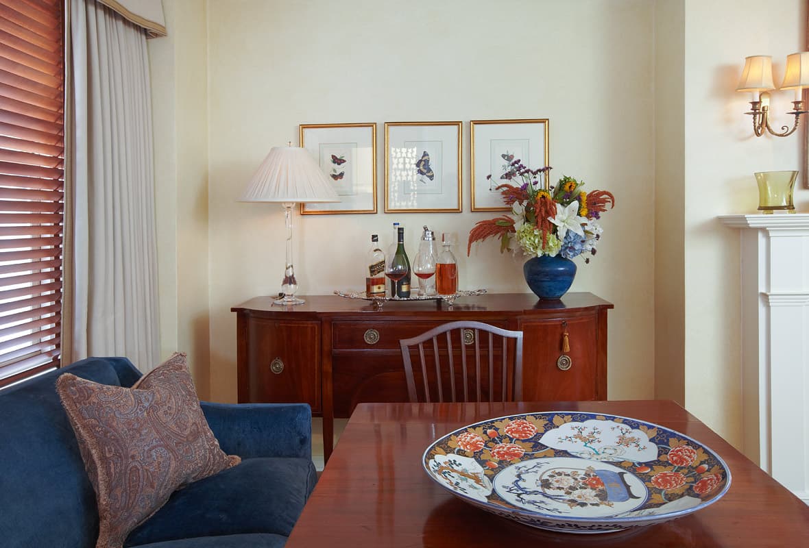 Mahogany sideboard under three gilt framed butterfly plates, decanters on a silver tray, market flowers in a blue stoneware vase, a blue velvet banquette and an Imari charger on the dining table
