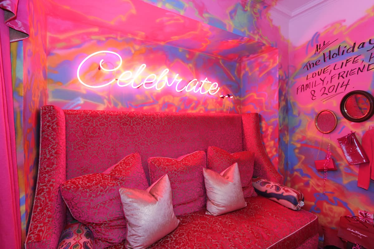 Red damask wing banquette piled with velvet cushions under a neon Celebrate script, hand sprayed mural walls, a marker dedication to love, life, family and friends, and round mirrors above lacquer boxes