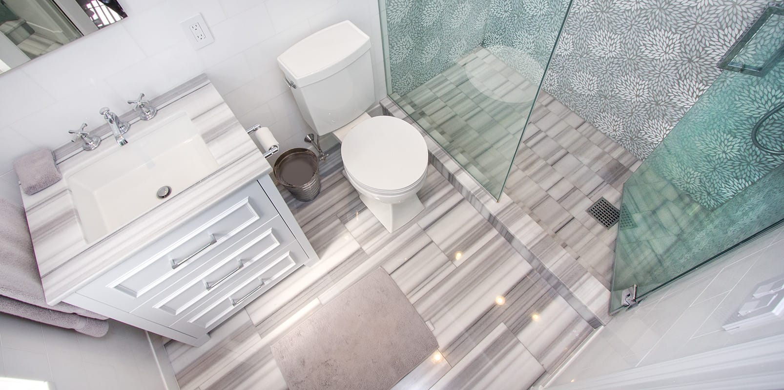 Bathroom seen from directly above, a white vanity in striated grey marble beside the toilet, a marble laid floor and a glass shower lined in aqua petal mosaic