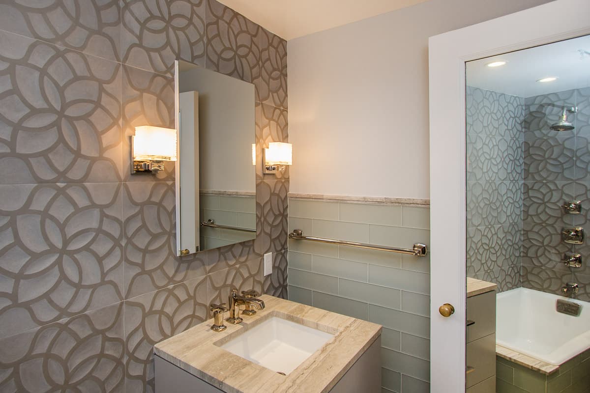Second bathroom vanity wall in large format petal patterned porcelain with a frameless mirrored cabinet between two glass sconces, sea glass tile below a marble cap rail and the tub and shower through the open door