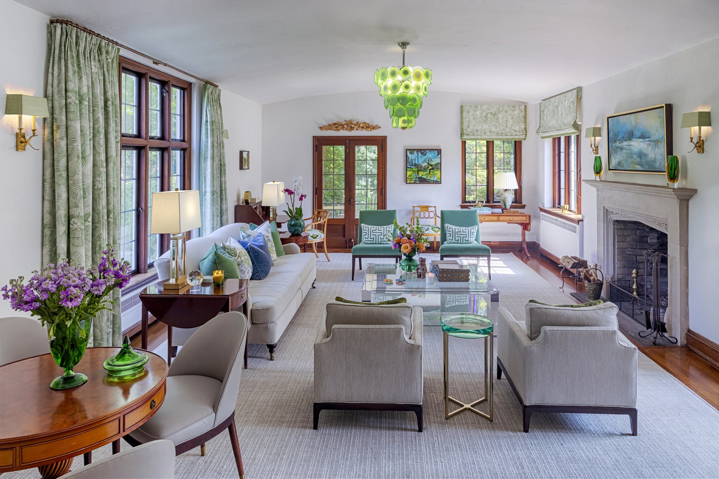 The living room along its length, a green glass disc chandelier under the barrel ceiling, cream sofas and club chairs on a pale carpet, floral curtains at the wood casements and a stone chimneypiece on the right